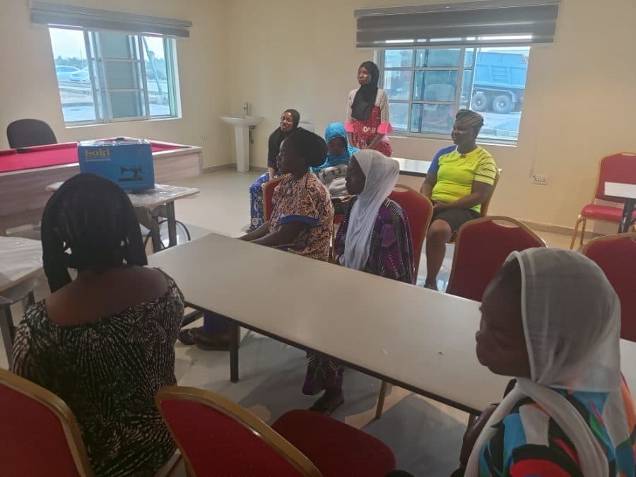 Participants seated around tables in the training room, a boxed sewing machine on the table beside them