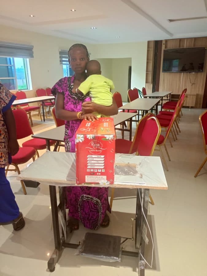 A woman holding her child stands beside the sewing machine she received at the empowerment programme