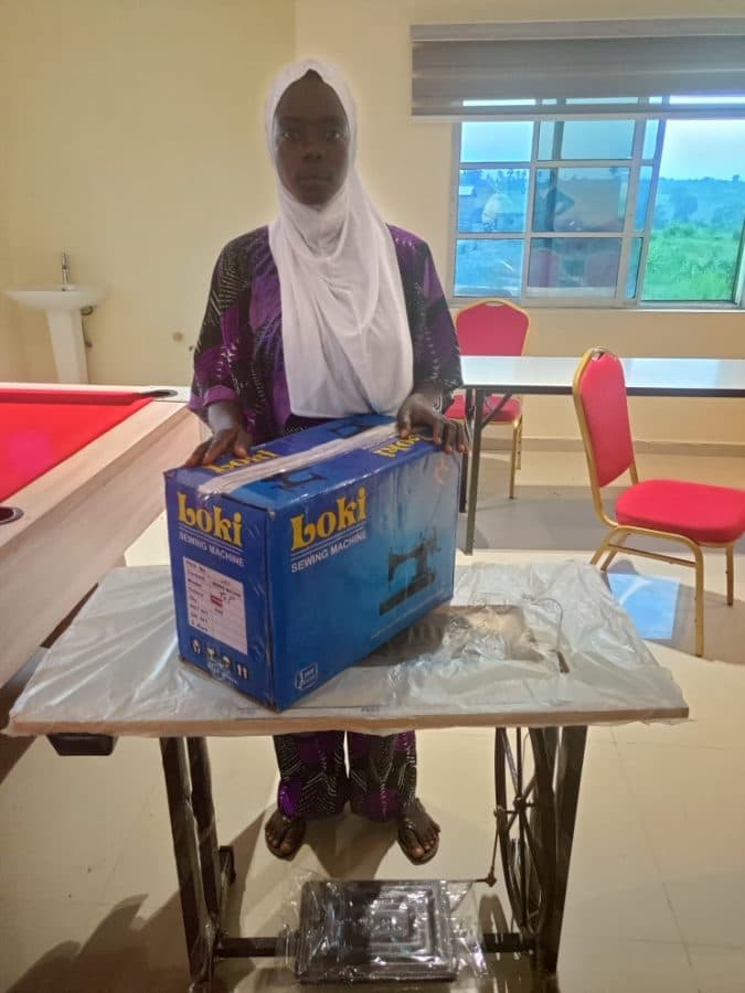 A participant in a white headscarf stands behind her boxed sewing machine and treadle stand